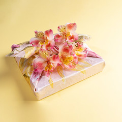 Gift box trendy wrapped in floral fabric in japanese Furoshiki technique with flowers Alstroemeria on yellow backdrop. Valentine's day, mother's day, Women's day or Birthday. Zero Waste. Square photo.