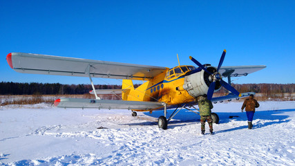 A yellow old biplane plane is parked on a winter airfield with technicians at work against a bright blue sky and white snow with the engine running and smoke
