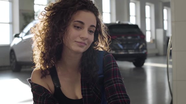 Close-up of gorgeous Caucasian woman with curly brunette hair and brown eyes sitting in sunlight and smiling. Beautiful young girl posing in sunrays in car repair shop. Gender equality, lifestyle.