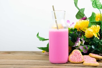 Obraz na płótnie Canvas Glass bottle filled with pink milk and a golden metal straw next to heart shaped cookies with pink frosting and sprinkles on wooden boards with a bouquet of flowers with a white background
