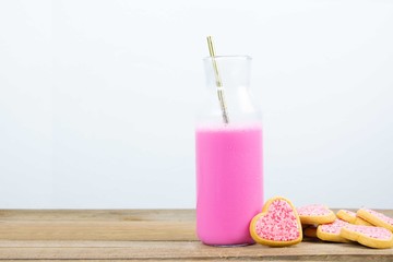 Glass bottle filled with pink milk and a golden metal straw next to heart shaped cookies with pink frosting and sprinkles on wooden boards and white background. Love concept