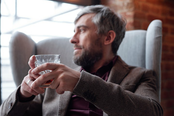 Close-up of introspective middle-aged man in jacket sitting in armchair in living room and enjoying...