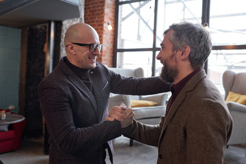 Cheerful bald man in glasses handshaking with friend while being excited to see him in own house