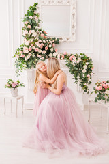 Fototapeta premium Blonde mom with cute daughter hugging. They are dressed in fancy dresses. Beautiful interior with flowers. The image of the princesses. The concept of the celebration on the birthday and women's day