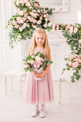 Beautiful blonde girl with long hair in a soft purple dress on a background of flowers posing. Cute baby model in the image of a Princess. She's wearing a full lilac dress
