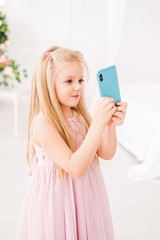 A beautiful blonde girl with long hair in a soft purple dress with a phone in her hands. Cute baby model takes photos on a smartphone camera.