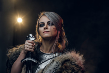 Portrait of a beautiful warrior woman holding a sword wearing steel cuirass and fur. Fantasy fashion. Studio photography on a dark background. Cosplayer as Ciri from The Witcher. - obrazy, fototapety, plakaty
