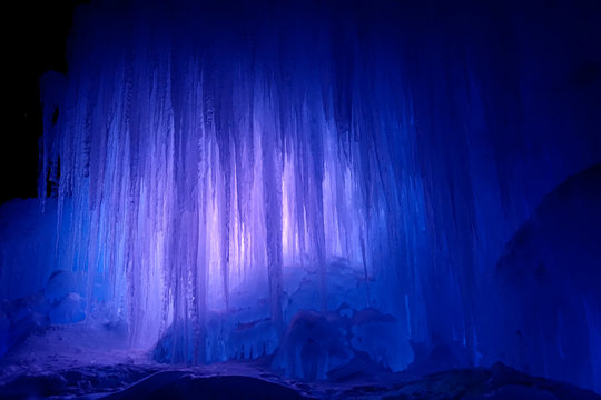 A massive wall of long ice stalactites are illuminated by a few rays of light.