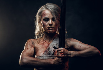 Naked Fantasy woman warrior wearing rag cloth stained with blood and mud posing with a sword. Studio photo on a dark background. Cosplayer as Ciri from The Witcher