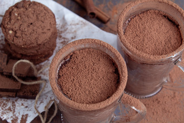 A pair of glass cups of hot chocolate with cinnamon on a wooden table with oatmeal cookies and chocolate