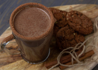 Transparent cup of hot chocolate with cinnamon and milk on a wooden board with oatmeal cookies and chocolate.