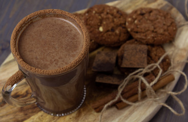 Transparent cup of hot chocolate with cinnamon and milk on a wooden board with oatmeal cookies and chocolate.