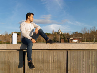 Young handsome caucasian man seated at Madrid Rio with the Almudena Cathedral behind him