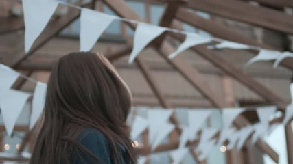 happy stylish young asian pretty girl in blue coat with wavy hair turns around, looks at camera, smiles on background of white festive triangular flags on rope hanging outdoors, swaying in wind