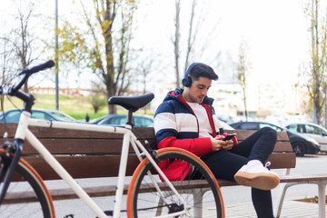Young man sitting on a bench in the street while using mobile phone and listening music by headphones