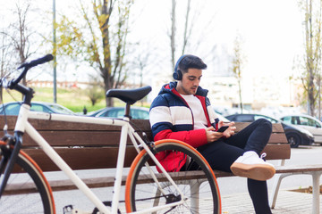 Young man sitting on a bench in the street while using mobile phone and listening music by headphones