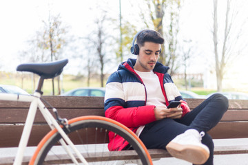 Obraz premium Young man sitting on a bench in the street while using mobile phone and listening music by headphones
