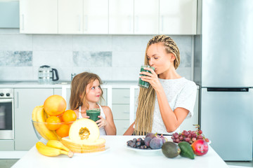 Happy woman mother and little girl daughter drinks green smoothie in kitchen. Mother and daughter with glass of natural detox smoothie in kitchen. Food supplements.