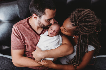 Multiracial parents kissing newborn baby wrapped in white blanket