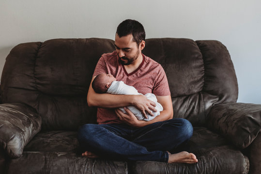Barefoot bearded dad on leather couch holding swaddled newborn