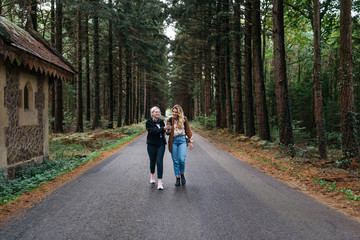 Two women walking on a road in the forest while watching their phone