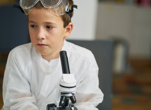 young boy looks at camera dressed in white coat prepares to observe by a microscope at home and do experiments