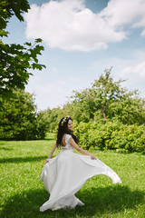 Young happy woman in white long dress has fun outdoors in a spring garden. Young bride with floral wreath in her wedding hairstyle spinning around and posing at nature in summer sunny day