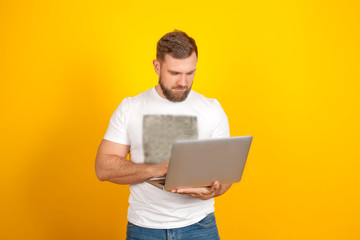 A man jokingly can't open a laptop, on a yellow background, in a white t-shirt and blue jeans, with copy space. Concept new modern technologies, gadgets and computer work.