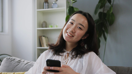 Close-up shooting of good-looking young Chinese woman in business clothes sitting on sofa and using smartphone. Fascinating girl looking at camera. Friendly face. Smile. Apartment.