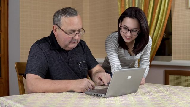 Daughter teaches father how to use a laptop. A young woman shows her old father where to click in a computer. The family is sitting in a comfortable room. A man looks at the camera and shows a thumb