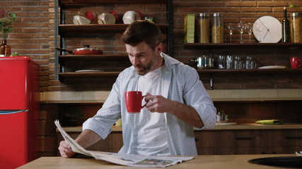 Serious middle-aged man having breakfast in the kitchen reading recent news on a paper magazine sitting by table at home.