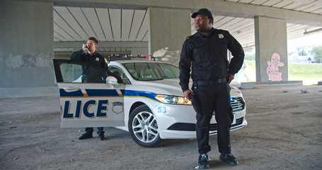 Young handsome policemen patrolling the city and protecting people. Afro american serious police officer standing by car while his colleague using walkie-talkie standing on door.