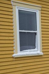 The exterior view of a double hung two pane glass window with a white mini blind. The window has beige trim around the white vinyl. The exterior wood wall of the building is a yellow in color.