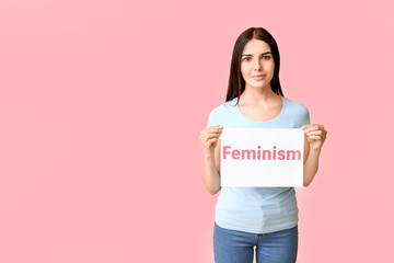 Young woman holding paper with text FEMINISM on color background