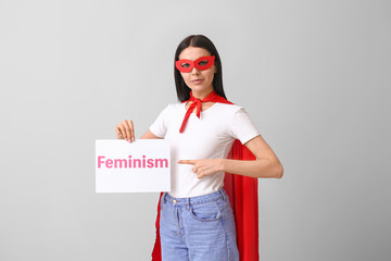 Young woman in superhero costume holding paper with text FEMINISM on grey background
