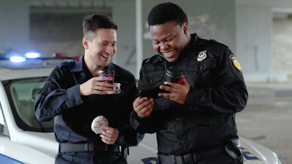 Cute young two policemen drinking coffee and eating donut use phone smile talk near patrol car help...
