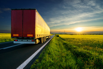 Red truck driving on the asphalt road between the yellow flowering rapeseed field in the rural landscape at sunset