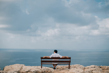 A man sits on a bench on the rocks overlooking the sea