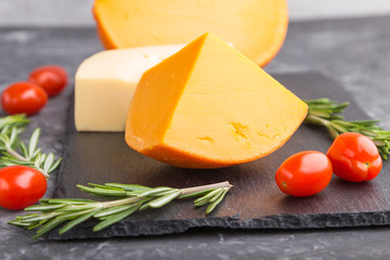 Various types of cheese with rosemary and tomatoes on black slate board on a black concrete background. Side view, close up.