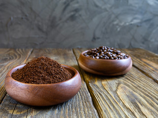 Ground coffee and coffee beans poured into wooden containers stand on a wooden table against a gray concrete wall.