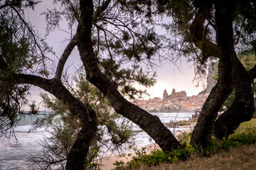 Seafront of Tyrrhenian Sea. View of the Sicilian city of Cefalu through trees growing on the shore