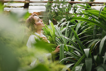 Young beautiful caucasian woman in glass greenhouse among colorful greenery leaves and flowers. Art portrait of a girl wearing a shirt. Gardener, nature lover, inspiration concept.