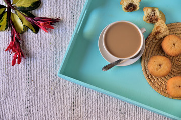 Breakfast in light green tray on woven blanket, cup with coffee and milk, cupcakes filled with sweet and red tropical flowers. Natural lighting, space to write.