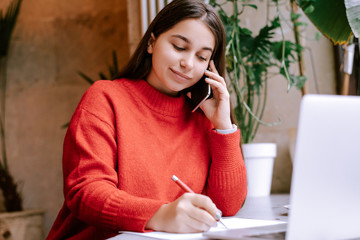 Young beautiful brunette woman  in red sweater making notes in notebook and talking on phone. Female freelancer connecting to internet via computer. Blogger or journalist writing new article 