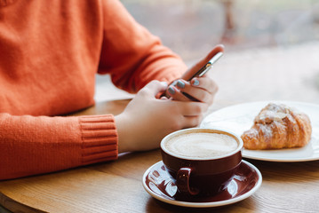 cropped view of girl holding smartphone near coffee and croissant on table
