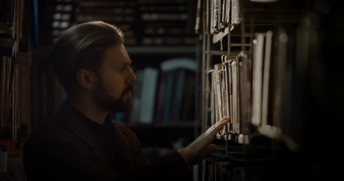 Handsome bearded male looking through books on library shelves in evening, closeup