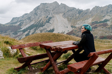 Fototapeta na wymiar A girl sits at a wooden table in the mountains near a cliff. A place for lunch with a gorgeous view