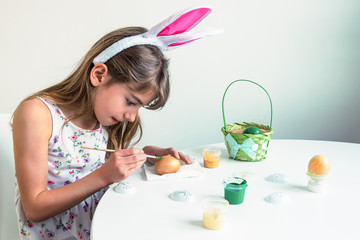 Cute little girl with bunny ears paints eggs at a white table. Easter concept.  Selective focus.