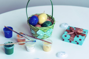 Basket with eggs, paints and a gift in a box on a white table. Easter concept.