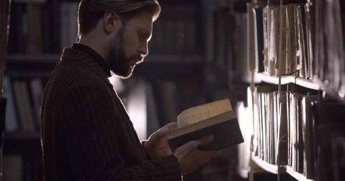 Handsome bearded hipster male turning pages in old dusty book in library storage, antique